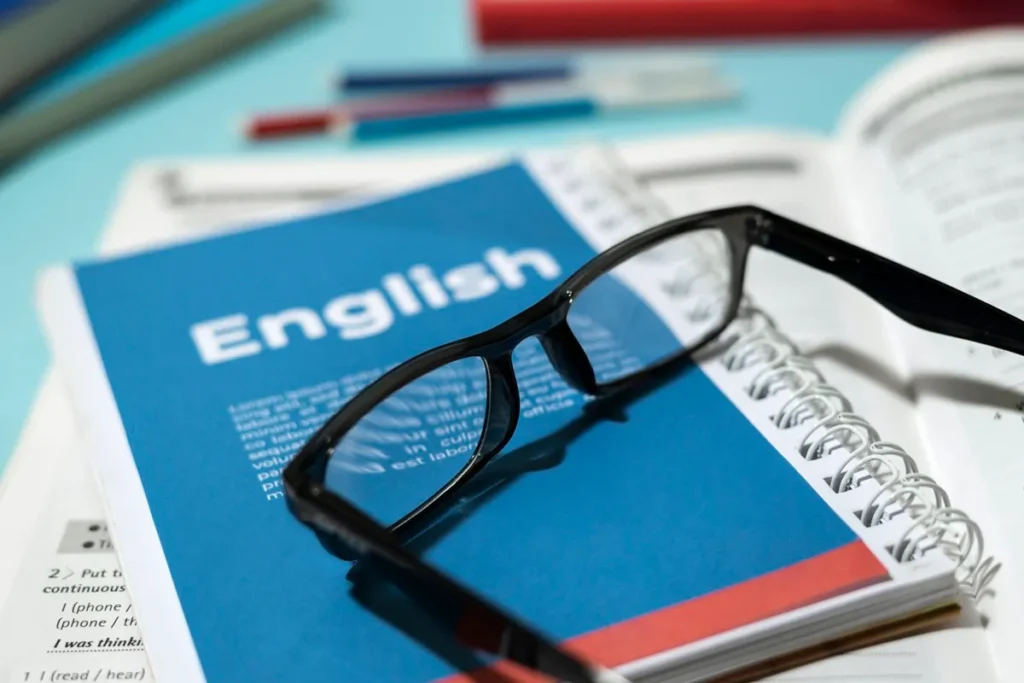 Caderno azul com a palavra "English" na capa, óculos de leitura e livros de inglês ao fundo, representando estudo do idioma.