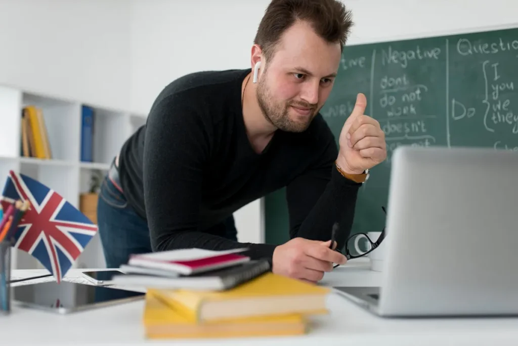 Homem com fones de ouvido fazendo gesto positivo em frente ao notebook, com lousa de gramática inglesa e bandeira do Reino Unido ao fundo.