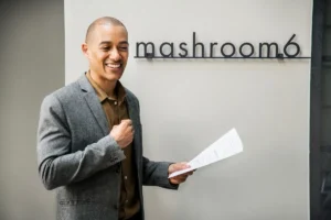 Homem sorridente usando blazer e camisa segurando folhas de papel, posicionado em frente a uma parede com a inscrição “mashroom 6”.