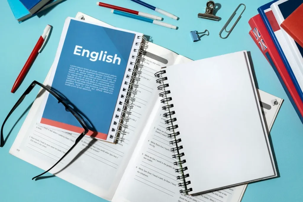 Livros e cadernos espiral com capa de inglês e exercícios gramaticais abertos sobre mesa azul, acompanhados de óculos, canetas e clipes de papel.