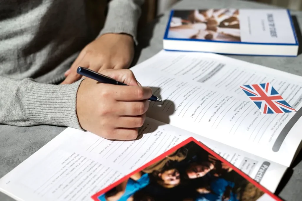 Mão de uma pessoa preenchendo exercícios em um livro didático de inglês com uma caneta, ao lado de uma bandeira do Reino Unido e outros materiais de estudo sobre uma mesa