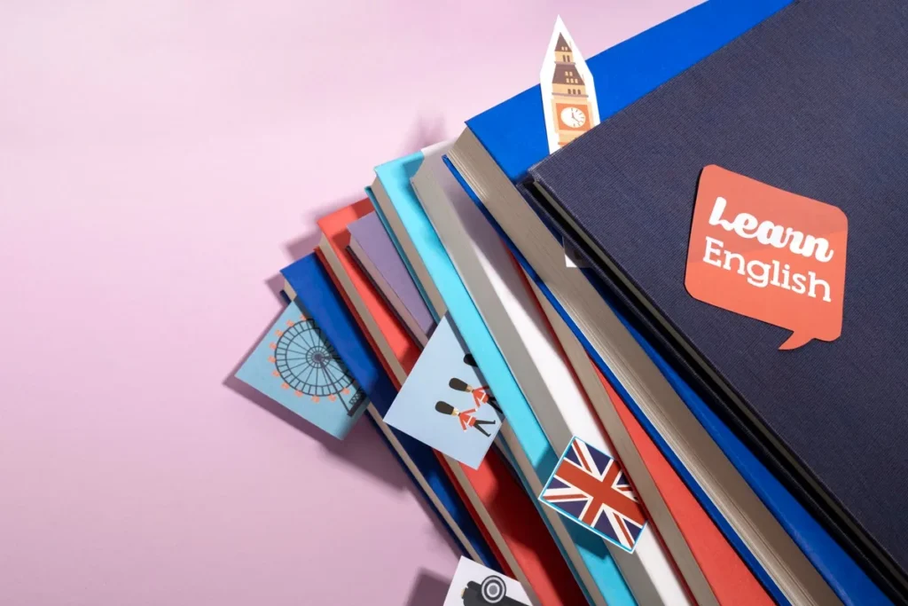 Vários livros empilhados com capa colorida e ilustrações do Reino Unido, como a roda gigante London Eye, guardas reais, Big Ben e bandeira britânica, sobre fundo rosa. Livro em destaque com etiqueta 'Learn English'.