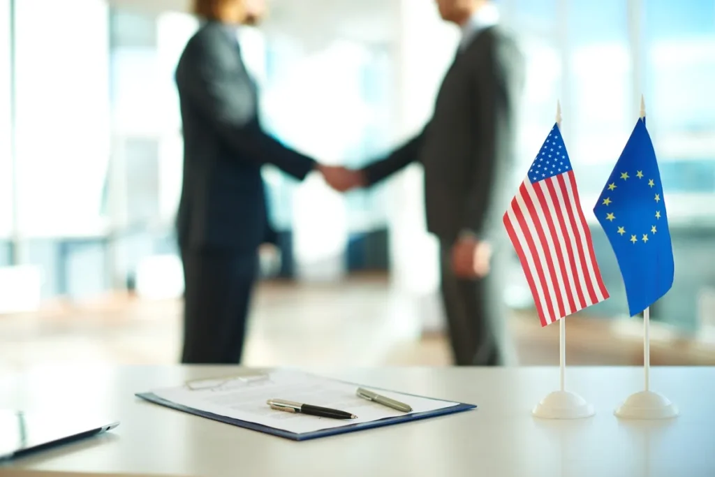 Bandeiras dos Estados Unidos e União Europeia sobre mesa com contrato e canetas, com dois empresários ao fundo apertando as mãos
