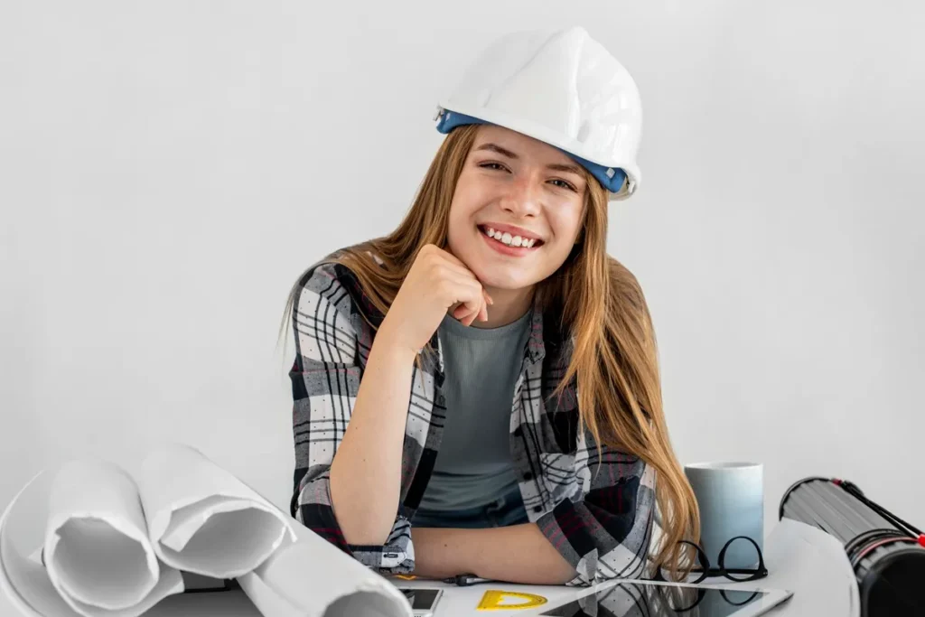 Engenheira sorridente com capacete branco, sentada à mesa com projetos arquitetônicos e instrumentos de medição.
