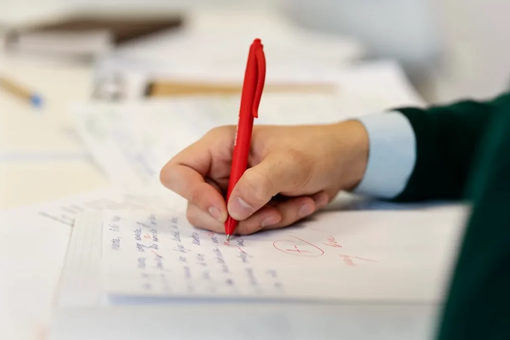 Pessoa corrigindo uma redação escrita em inglês com caneta vermelha sobre folha de caderno, destacando anotações e observações no texto.