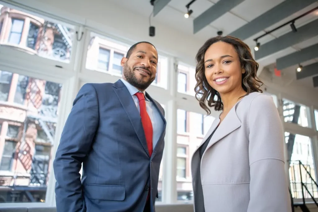 Um homem e uma mulher, ambos profissionais C-level, sorriem para a câmera em um ambiente corporativo moderno e bem iluminado. O homem veste um terno azul marinho e gravata vermelha, enquanto a mulher usa um blazer cinza claro. A imagem transmite confiança, sucesso e liderança no mundo corporativo.