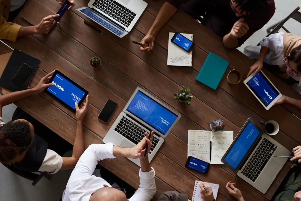 Vista superior de uma reunião de executivos C-Level em uma mesa de madeira. Os profissionais utilizam notebooks, tablets e smartphones, enquanto discutem estratégias de negócio e colaboram em um ambiente corporativo.