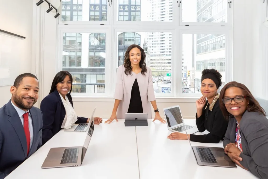 Uma equipe diversificada de executivos C-level sorri para a câmera durante uma reunião de negócios. Uma mulher no centro, em pé, lidera a equipe. A imagem transmite colaboração, sucesso e liderança executiva.