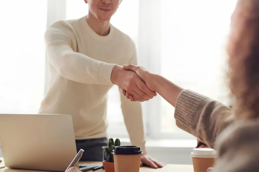 Dois profissionais C-level apertam as mãos sobre uma mesa de escritório com laptops e copos de café. A imagem simboliza acordo, parceria e sucesso em nível executivo.