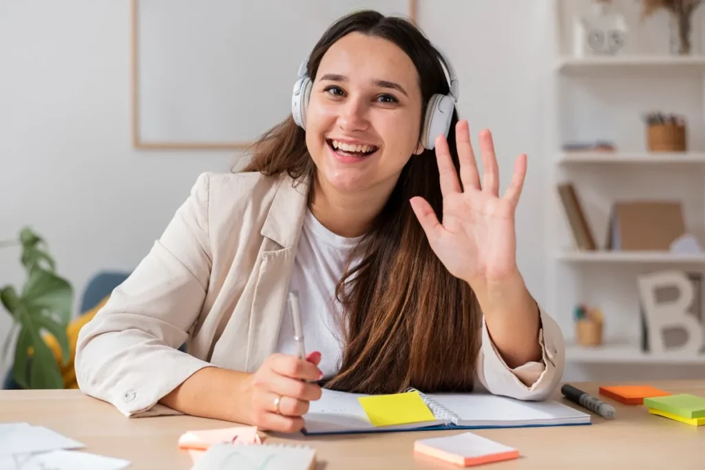 Jovem acenando e sorrindo enquanto participa de uma videoconferência, usando fones de ouvido e sentada em mesa com materiais de escritório