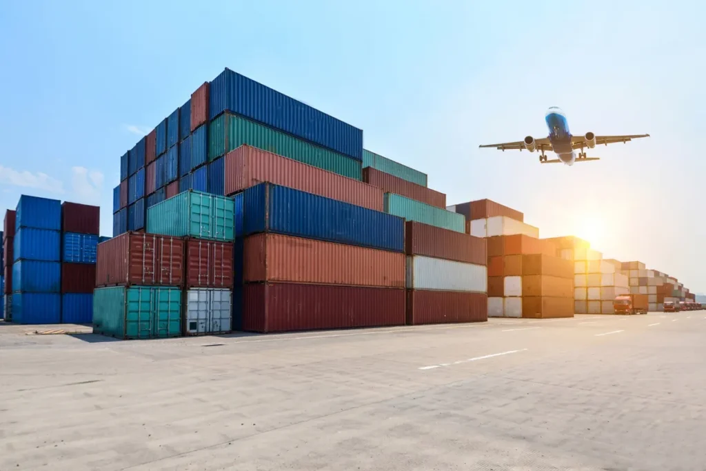 Um pátio de contêineres coloridos com um avião voando sobre eles. A imagem destaca a logística, o transporte e o comércio exterior.
