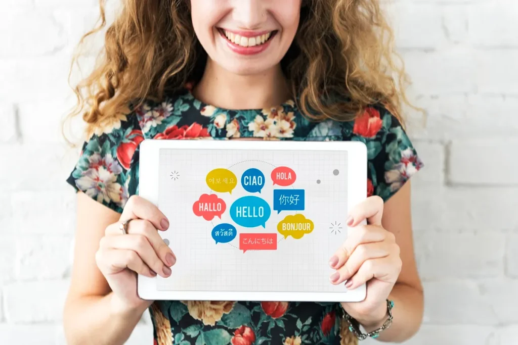 Imagem de uma mulher sorrindo e segurando um tablet, exibindo balões de fala coloridos com saudações em diversos idiomas, como "HELLO", "HOLA", "BONJOUR", entre outros.