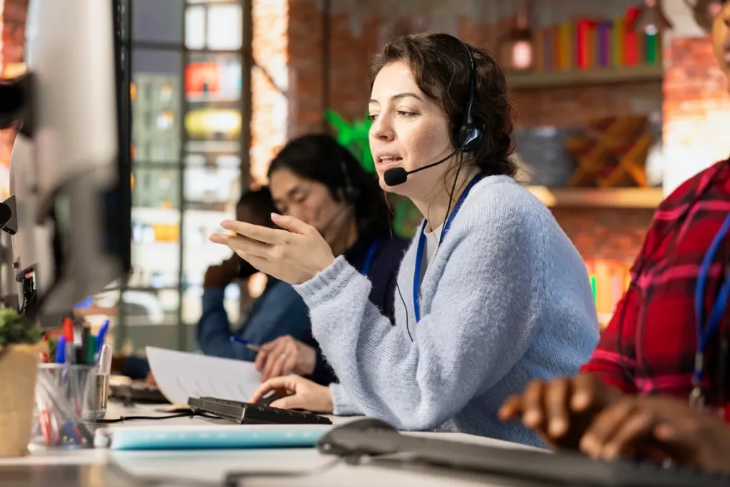 Mulher trabalhando em um escritório de atendimento ao cliente, usando headset e gesticulando enquanto fala, com outros colegas ao fundo.
