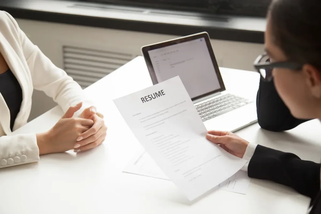 Imagem de uma entrevista de emprego com um recrutador analisando o currículo de um candidato, com a palavra 'RESUME' em destaque na parte superior do documento. Um laptop está visível ao fundo, com um ambiente de escritório profissional