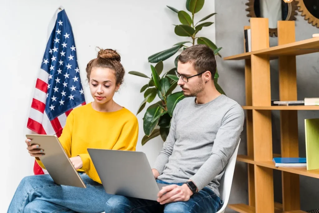 Casal estudando inglês, com uma mulher usando tablet e um homem com laptop, com a bandeira dos EUA ao fundo
