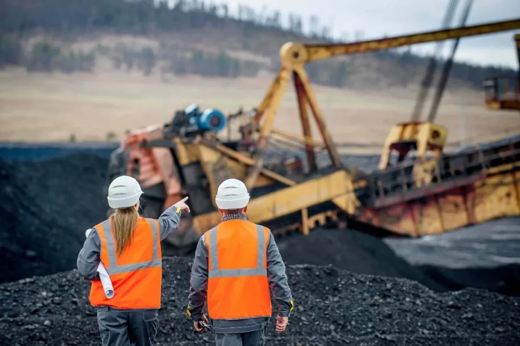 Dois engenheiros de mineração com capacetes e coletes de segurança, inspecionando uma escavadeira gigante em uma mina.