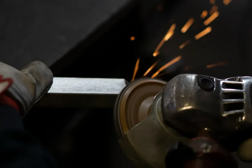 Trabalhador com luvas usando uma lixadeira em uma barra de metal, criando faíscas.