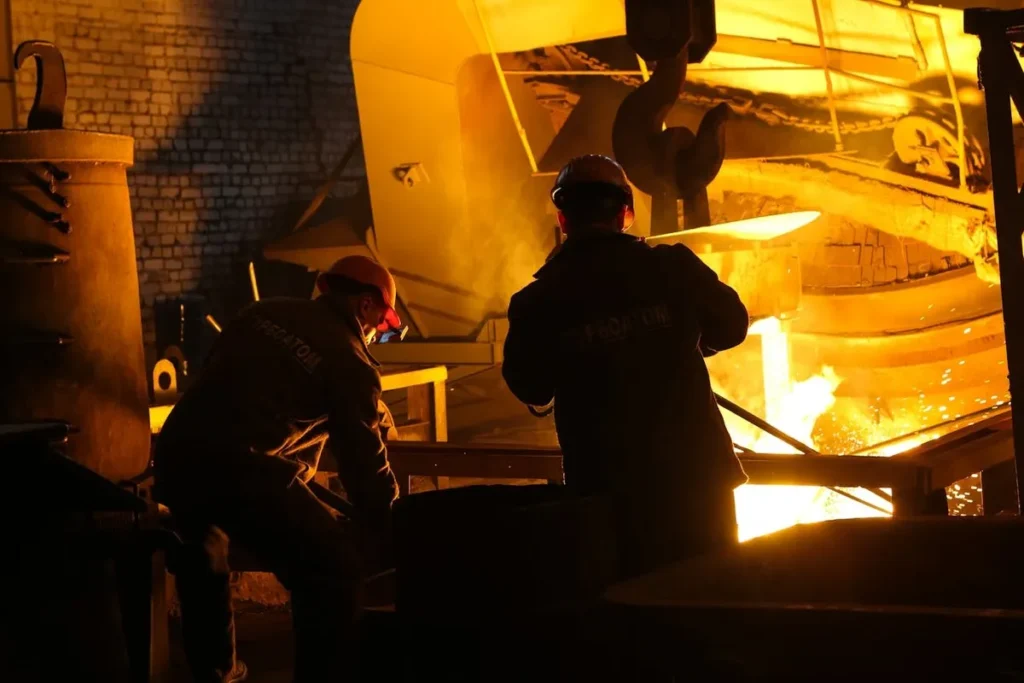 Dois operários em uma siderúrgica com forno incandescente e metal derretido.