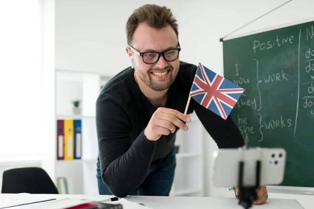 Professor de inglês sorrindo e segurando uma bandeira do Reino Unido, enquanto ensina com um quadro-negro ao fundo e um celular gravando