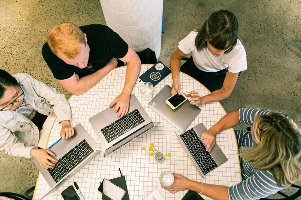 Uma vista aérea de quatro membros de uma equipe de startups em torno de uma mesa redonda, trabalhando em laptops. A imagem representa o ambiente colaborativo e a produtividade de uma startup.