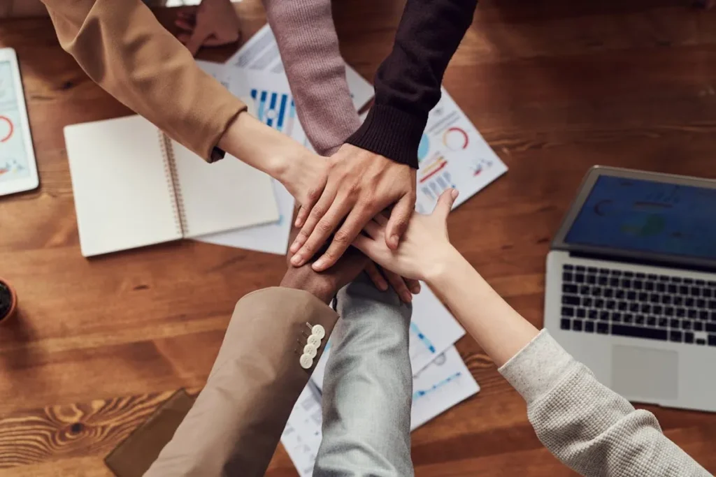 Mãos de um grupo diverso de profissionais de startups se unem no centro, sobre uma mesa cheia de documentos e um laptop. A imagem representa trabalho em equipe, união e sucesso em uma startup.