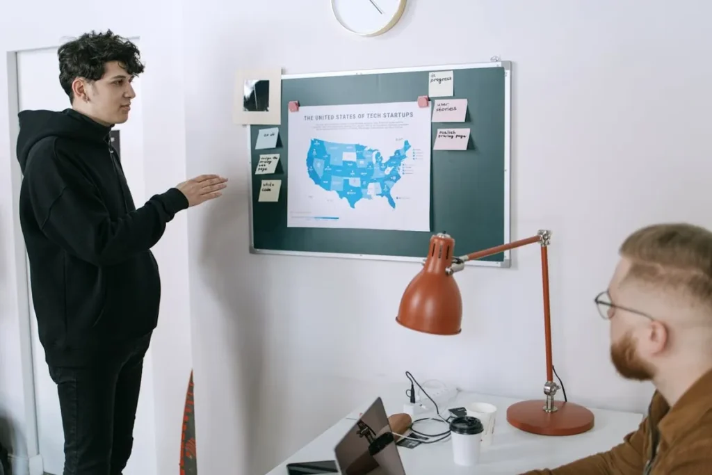 Um jovem empreendedor apresenta dados sobre startups em um mapa dos EUA para um colega em um escritório minimalista. A imagem representa estratégia, planejamento e o ambiente de trabalho em uma startup.