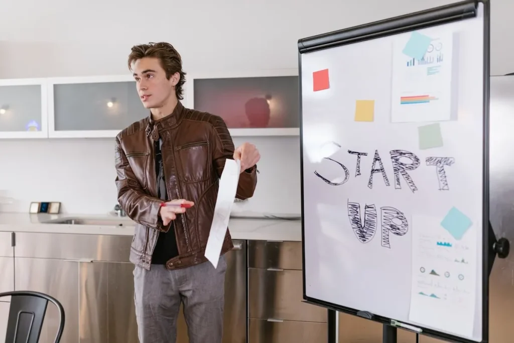 Jovem empreendedor em pé, apresentando uma ideia sobre startups em um quadro branco. A imagem representa inovação, criatividade e o ambiente de trabalho em uma startup.