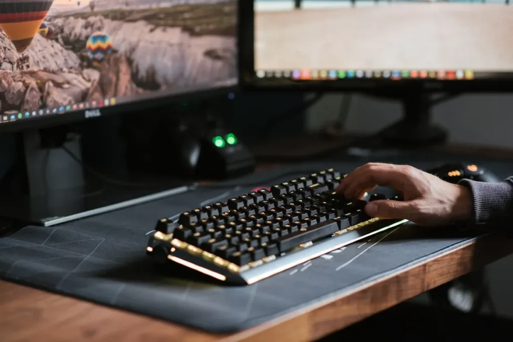 Uma pessoa usando um teclado de computador em uma mesa com dois monitores. A imagem representa o trabalho na área de TI (Tecnologia da Informação) e o ambiente de trabalho de um profissional de tecnologia.