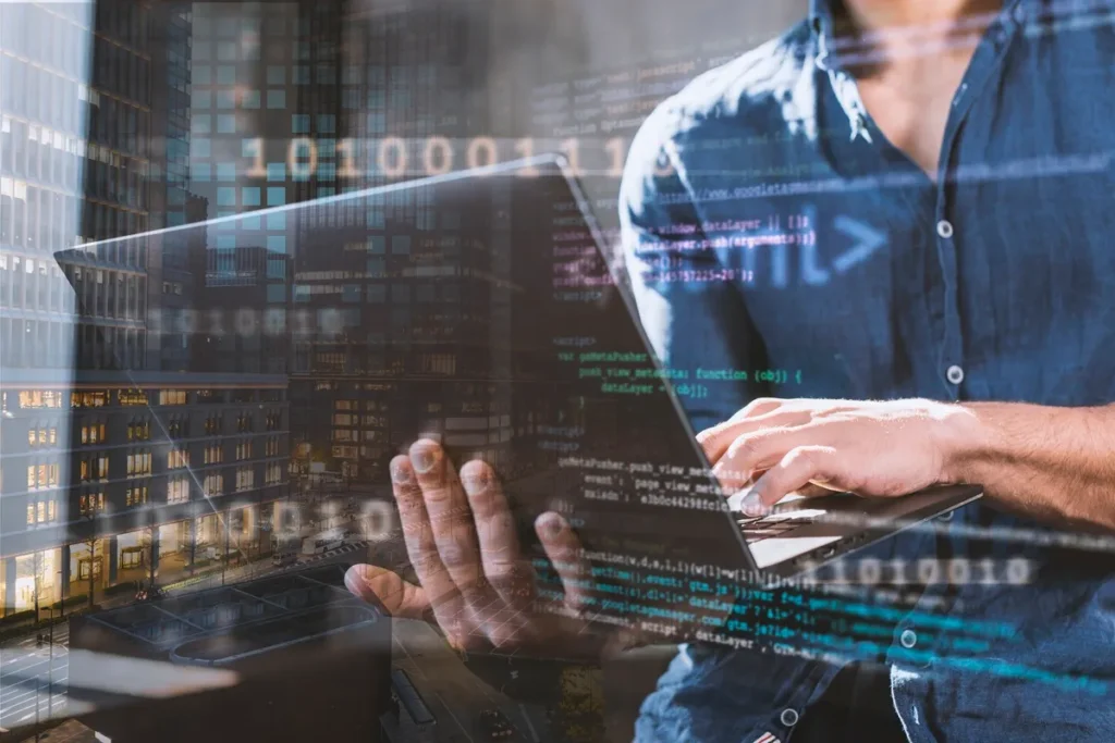 Programador de camisa azul digitando em um laptop enquanto código binário e informações digitais são exibidas ao fundo, com vista para prédios urbanos.