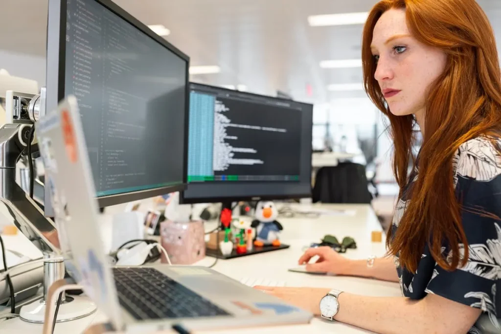 Uma programadora com cabelo ruivo e sardas trabalhando com dois monitores e um laptop. A imagem destaca a importância da programação e da TI (Tecnologia da Informação).
