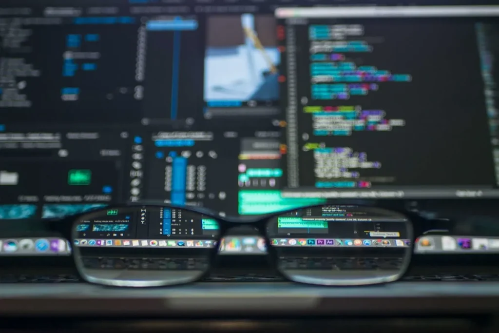 Um par de óculos sobre a mesa com a reflexão de um monitor de computador exibindo códigos de programação, software de edição e dados. A imagem destaca a complexidade e a importância da TI (Tecnologia da Informação) no dia a dia.