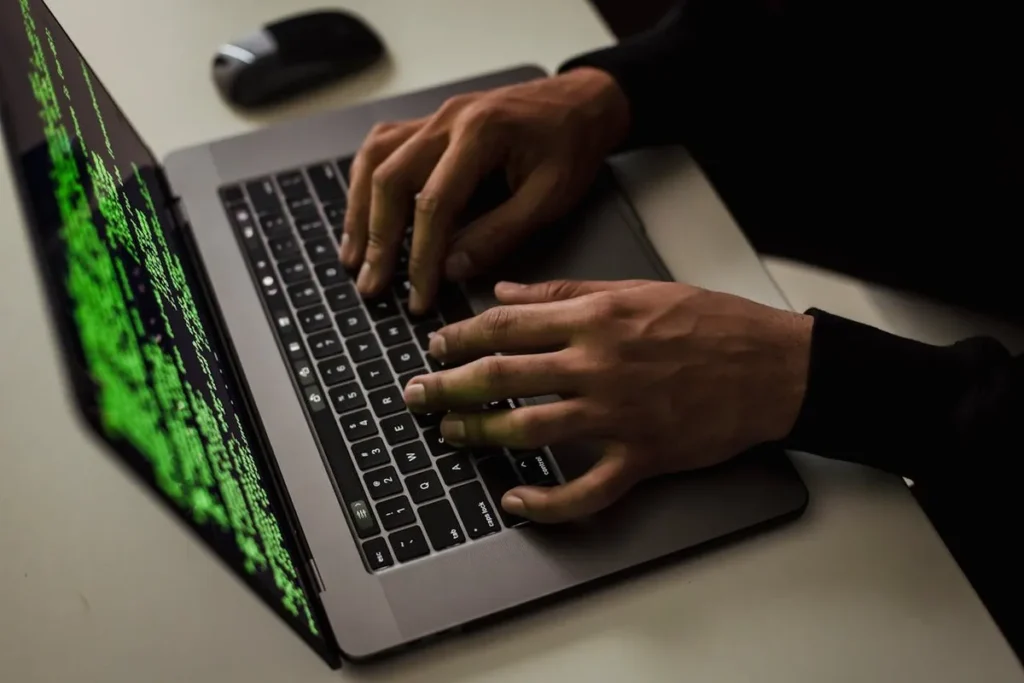 Pessoa digitando em um notebook com códigos verdes de segurança cibernética na tela. A imagem representa o trabalho em TI (Tecnologia da Informação) e o hacker ético.