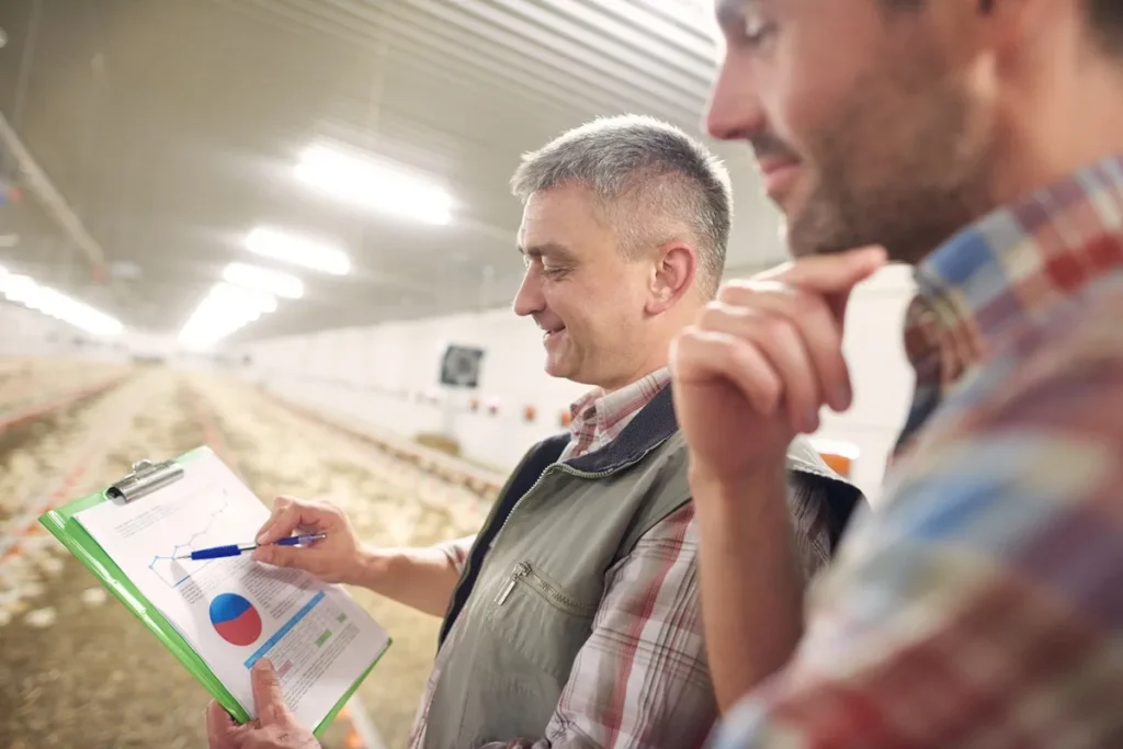 Dois profissionais do setor agropecuário observando gráficos de desempenho e produtividade em um ambiente de granja, com um gráfico de linhas e gráfico de pizza no clipboard.