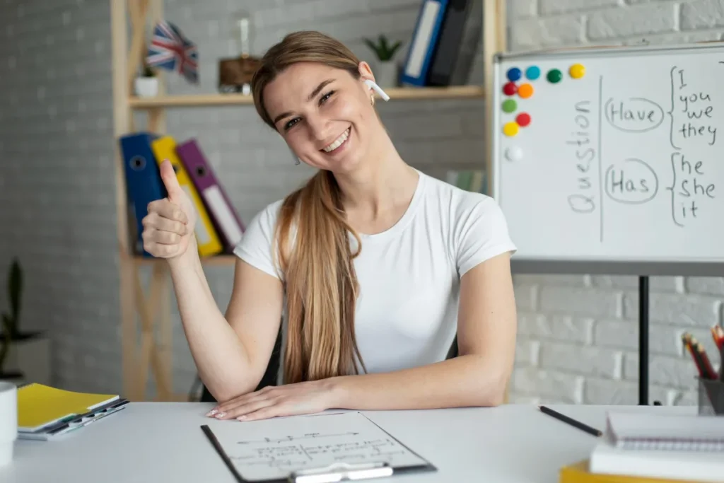Professora sorridente com fone de ouvido e lousa com lição de inglês ao fundo