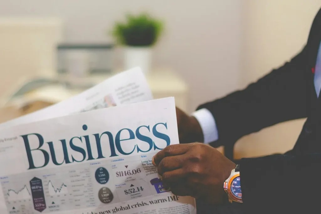 Homem de terno lendo um jornal com título 'Business' e gráficos financeiros visíveis