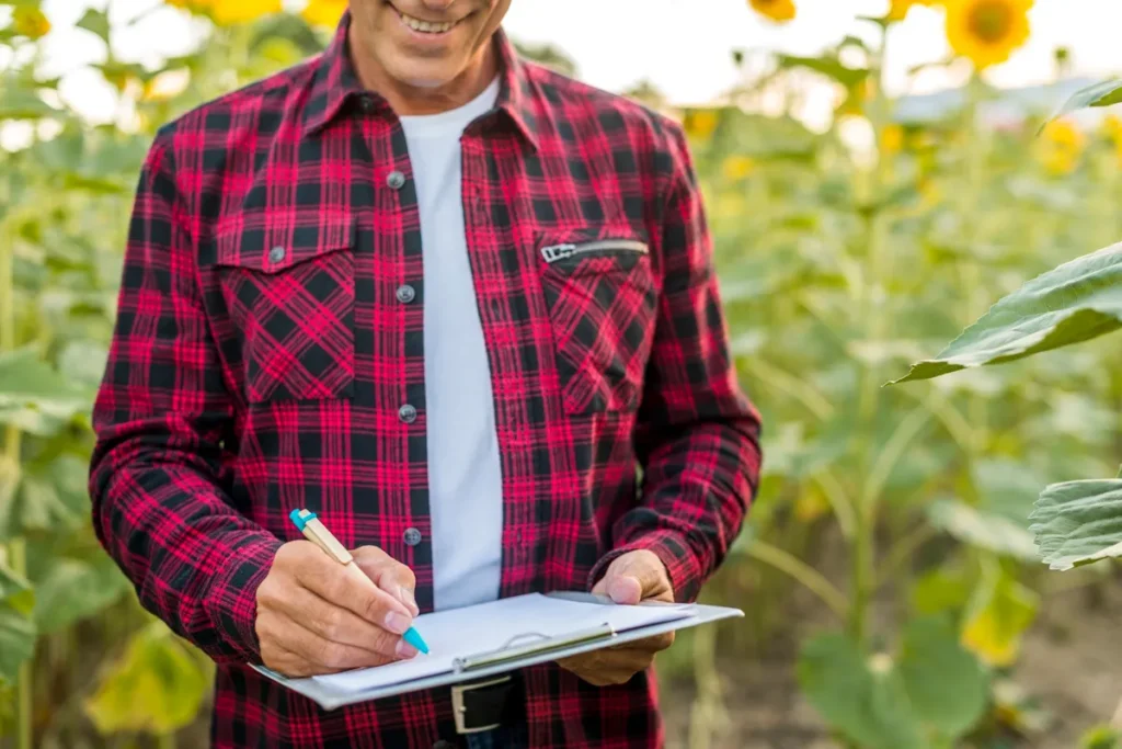 Homem em campo de girassóis anotando dados em um clipboard, com camisa xadrez vermelha, observando atentamente as informações.