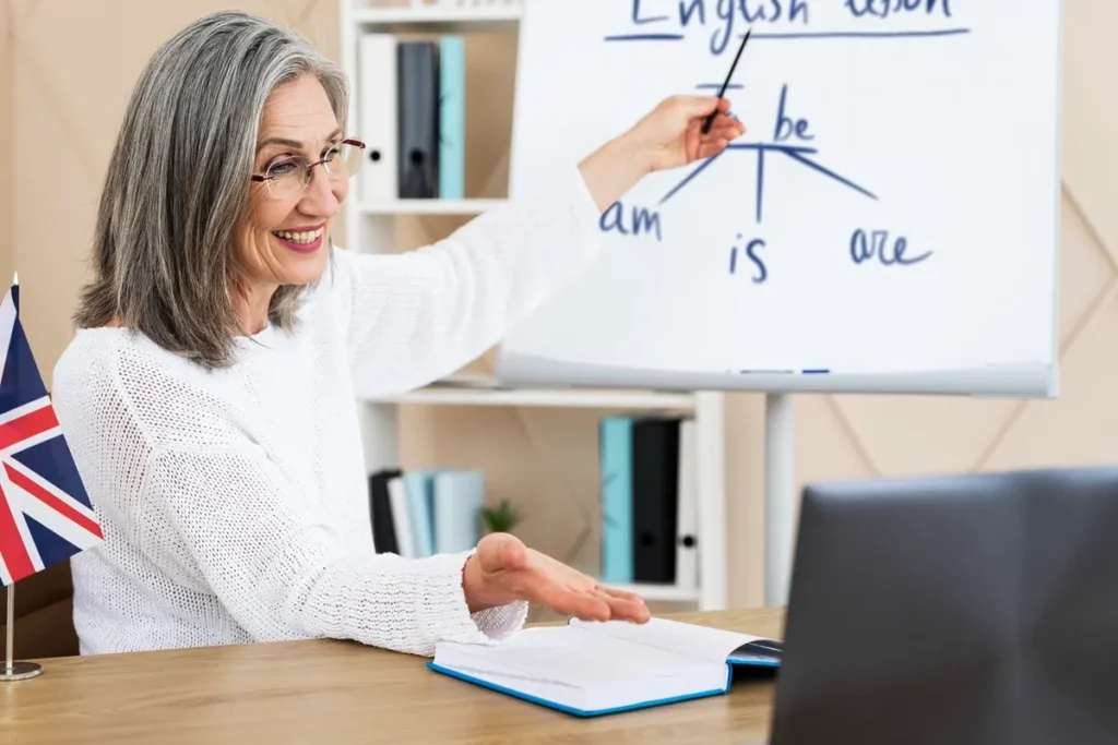 Professora idosa sorridente ensinando o verbo "to be" em um quadro branco durante aula de inglês, com bandeira do Reino Unido ao fundo