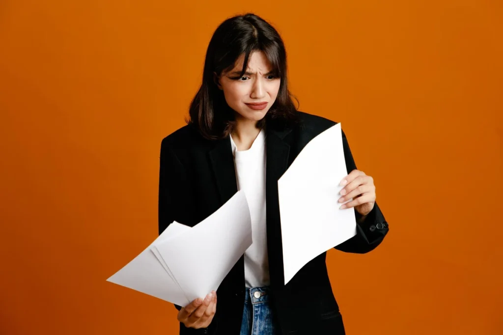 Mulher com expressão confusa analisando documentos em fundo laranja, representando frustração ou reclamações profissionais.