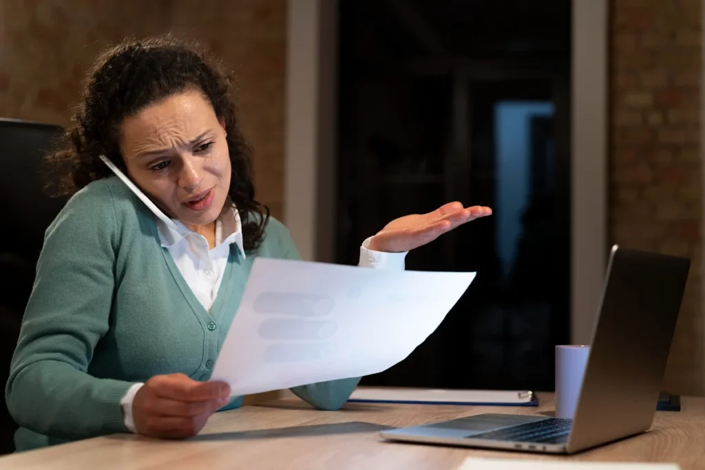 Mulher estressada ao telefone segurando um documento, sentada à mesa com um laptop, demonstrando frustração durante uma chamada de trabalho.
