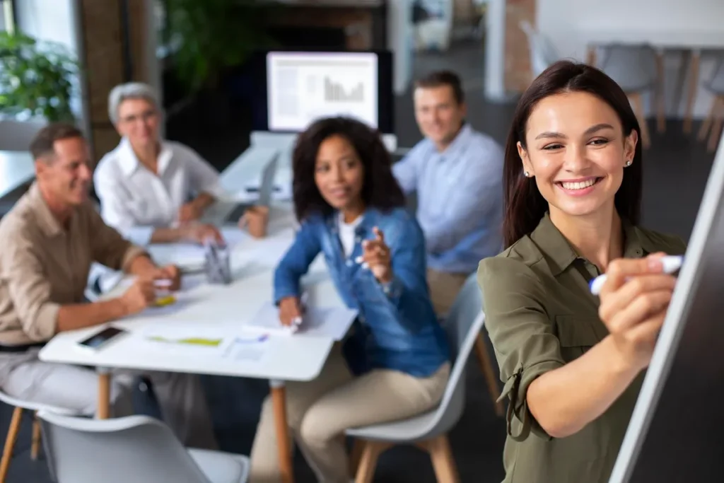 Profissional feminina sorrindo enquanto escreve em um quadro branco, com outros membros da equipe ao fundo em uma reunião de trabalho.