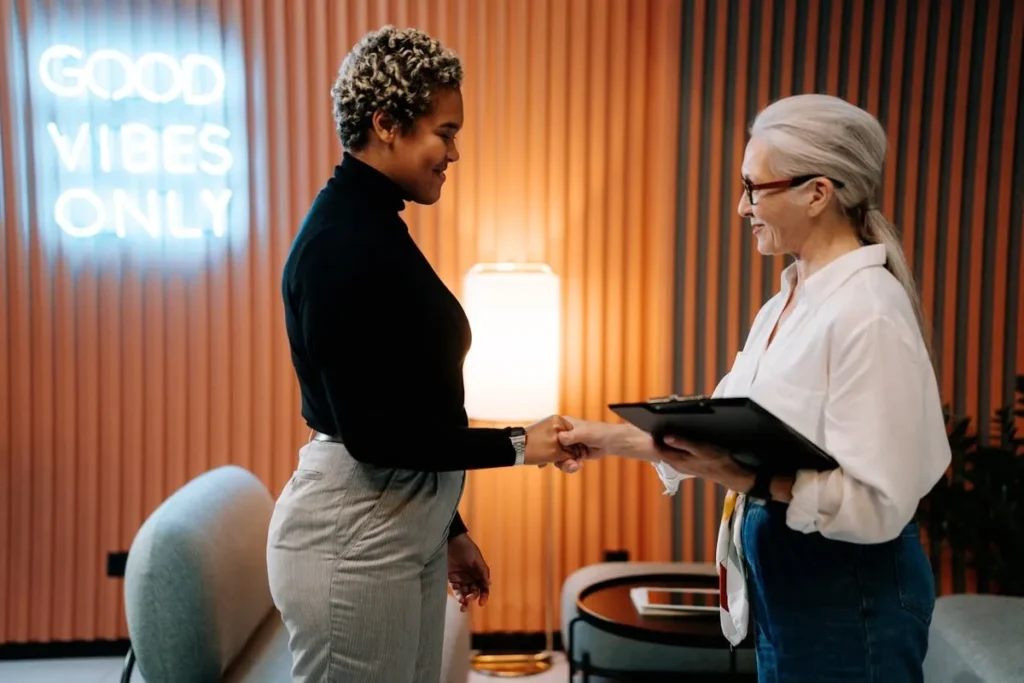 Duas mulheres, uma recrutadora e uma candidata, apertando as mãos em uma entrevista de recursos humanos.