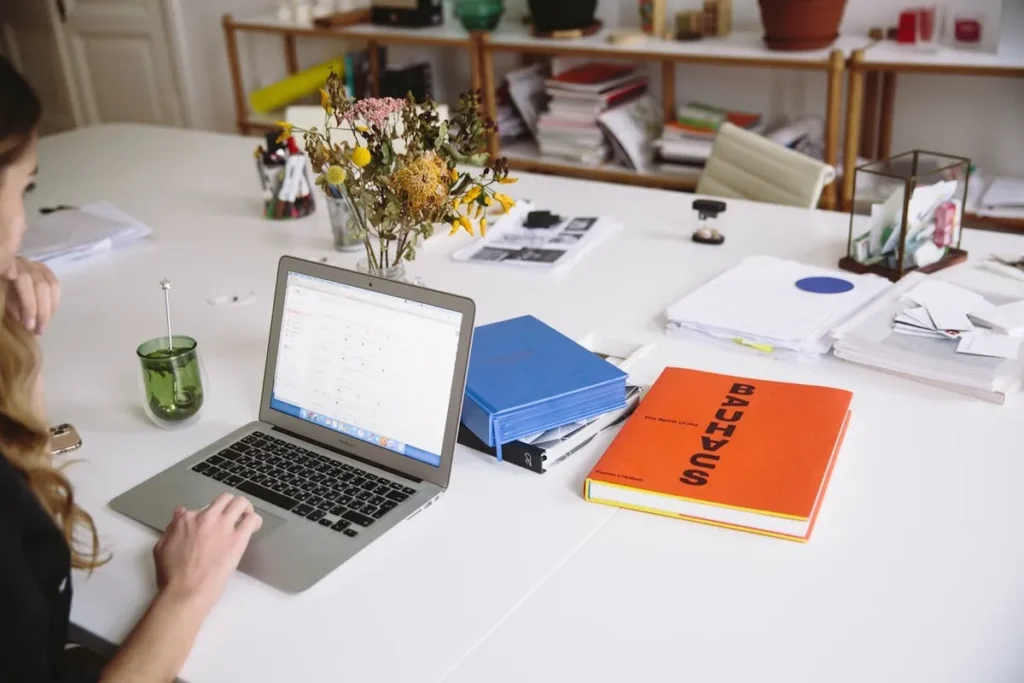 Mulher trabalhando em laptop com livros e documentos, simbolizando a gestão de recursos humanos.