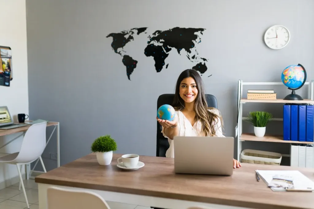 Mulher sorridente segurando um globo terrestre em um escritório moderno, com mapa-múndi na parede e elementos de decoração relacionados a viagens.