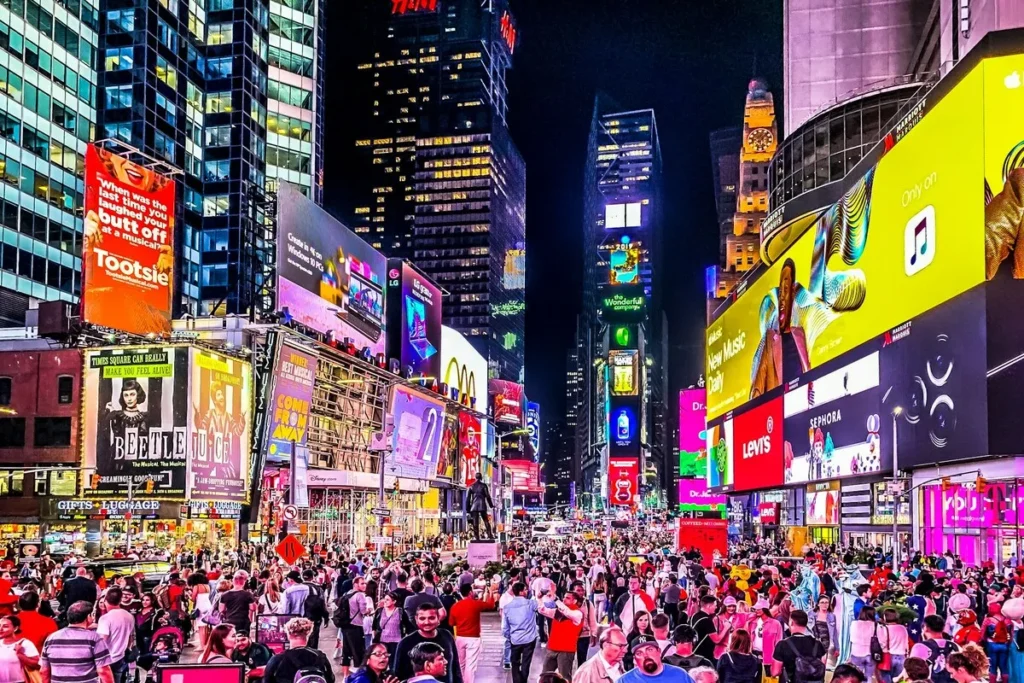 Multidão de turistas na Times Square à noite, cercada por letreiros luminosos e outdoors coloridos em Nova York.