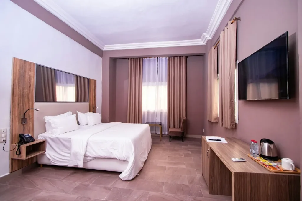 Quarto de hotel moderno e aconchegante com cama de casal, cortinas bege, TV de tela plana e mesa com chaleira elétrica e garrafas de água.
