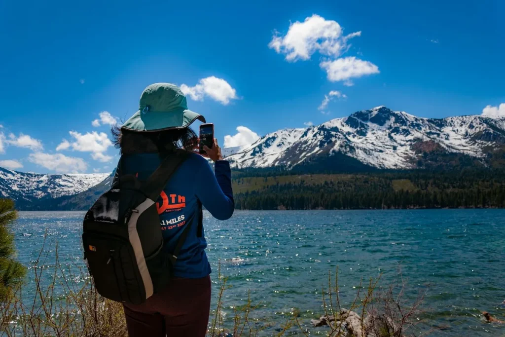 Mulher com mochila tirando foto de montanhas cobertas de neve ao lado de um lago azul