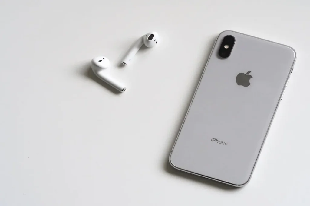  Um smartphone iPhone prateado com a parte traseira virada para cima e um par de fones de ouvido sem fio brancos (AirPods) ao lado, ambos sobre uma superfície branca lisa.
