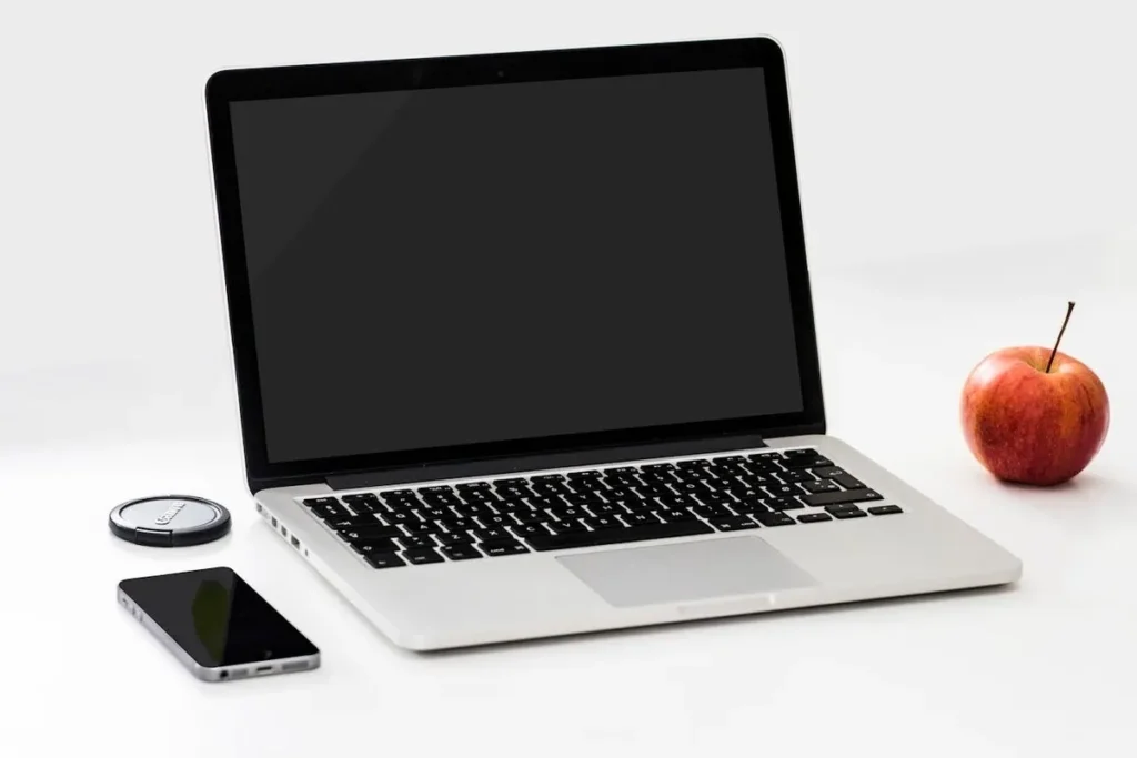 Um notebook prateado com a tela preta vazia, um smartphone preto ao lado esquerdo e uma maçã vermelha à direita, todos sobre uma superfície branca, sugerindo um ambiente de trabalho minimalista.