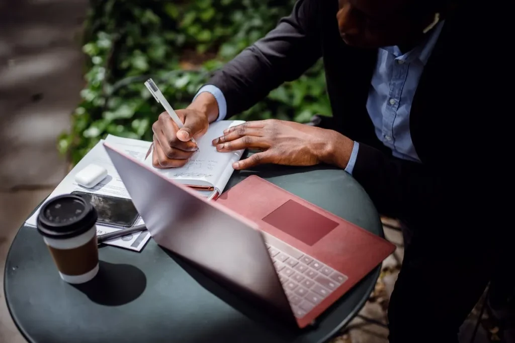Um homem de pele escura vestindo um terno preto e camisa azul, escrevendo em um caderno aberto sobre uma mesa redonda ao ar livre, com um laptop rosa aberto, um copo de café, um smartphone e um estojo de fones de ouvido ao lado.