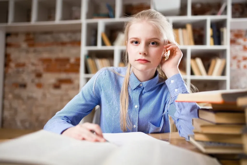 Uma jovem aluna de cabelo loiro sentada em uma mesa com livros de inglês abertos, concentrada em seus estudos. Ao fundo, uma estante com livros e uma parede de tijolos.
